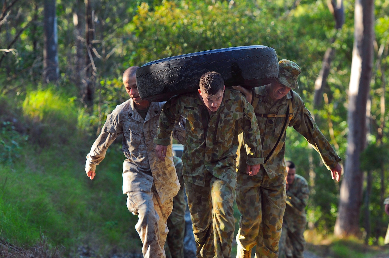 Three men in military uniforms carry a large tire.