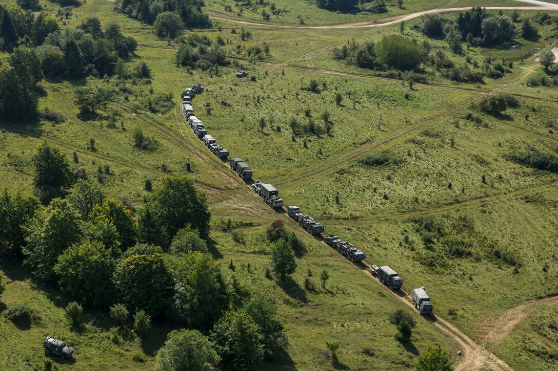 Army vehicles seen from overhead drive on a road through green terrain.