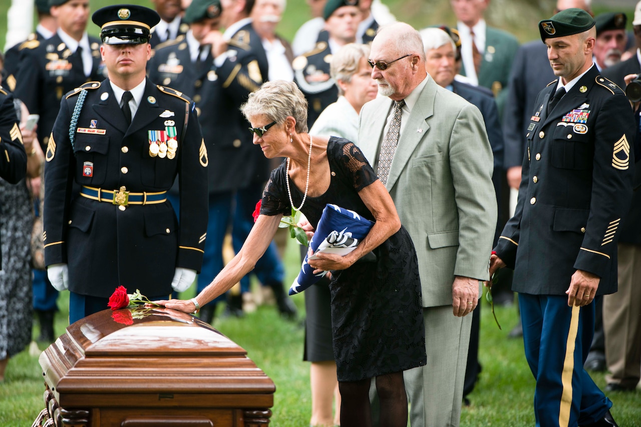 A woman places a rose on a casket in a cemetery with other people in the background.