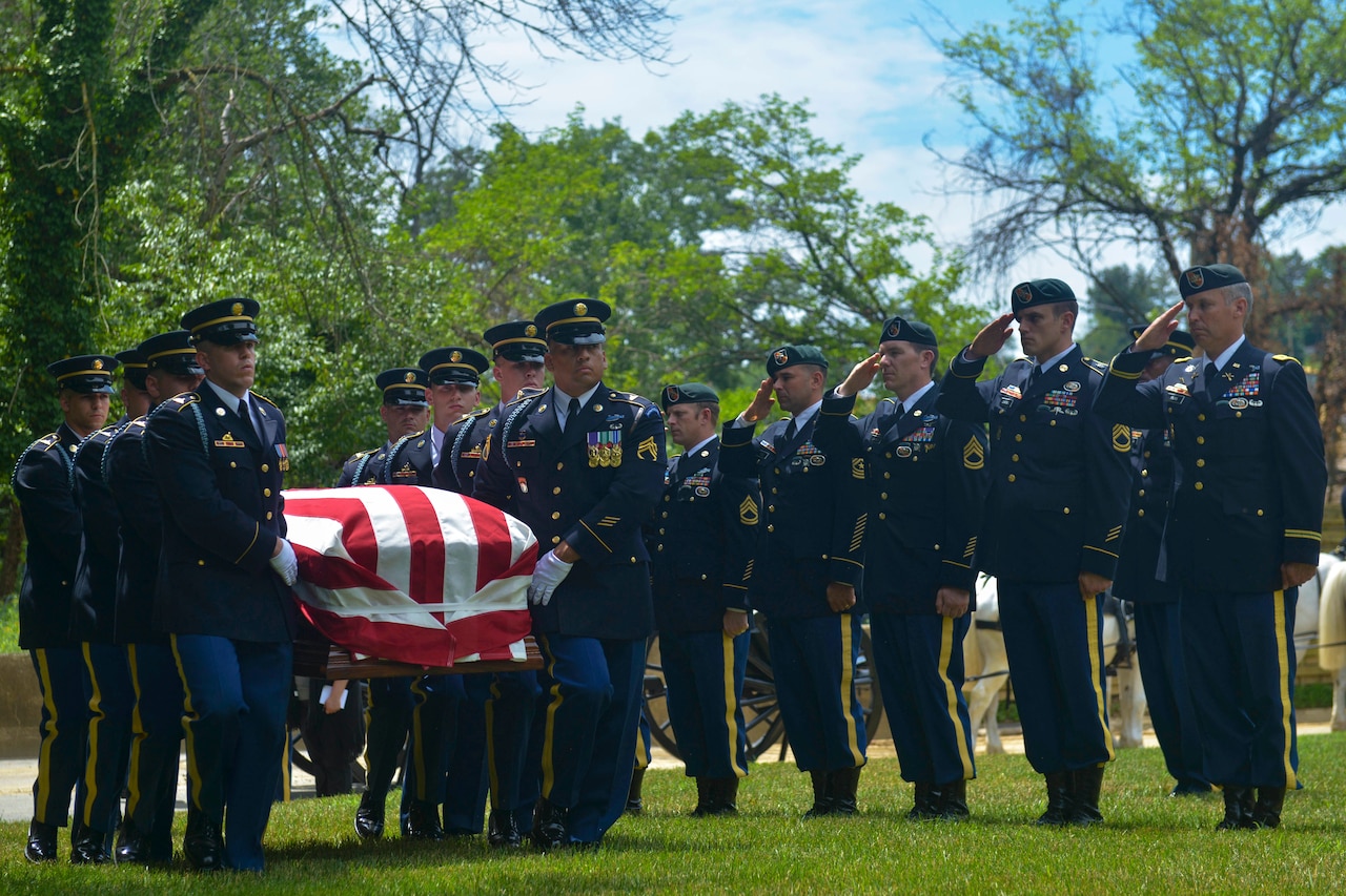 A group of soldiers salute while others carry a casket in a cemetery.