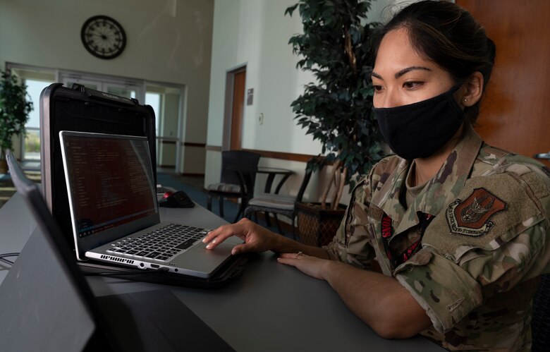 A woman wearing a facemask at a desk looks at a laptop.