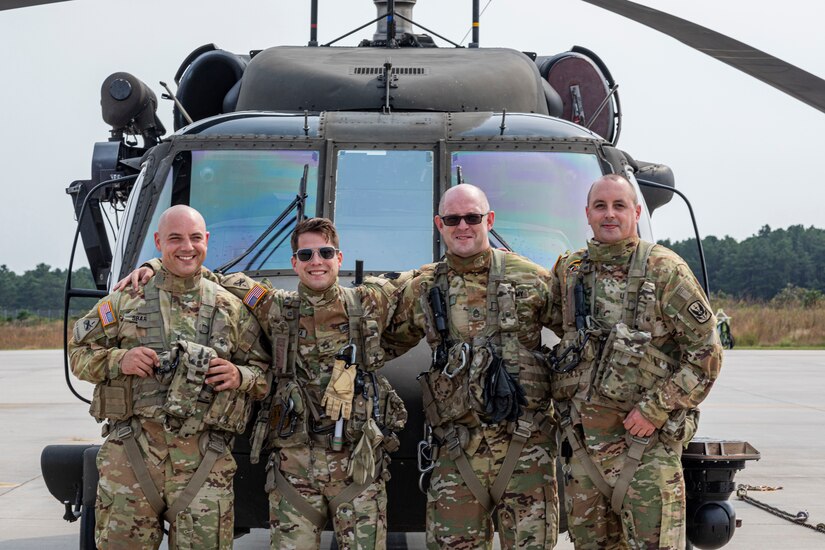 Four soldiers smile as they stand in front of a helicopter.