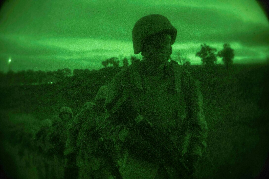 Marines line up while carrying weapons in a field as through night vision goggles.