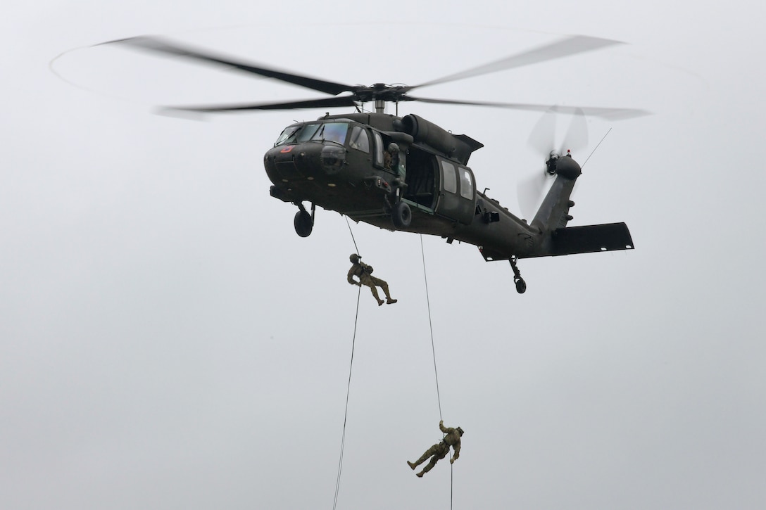 Soldiers rappel down a rope attached to an airborne helicopter.