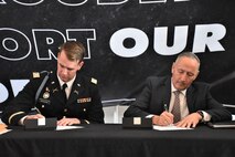 two men sitting at a table, one in a suit and the other in an Army uniform, signing a piece of paper.