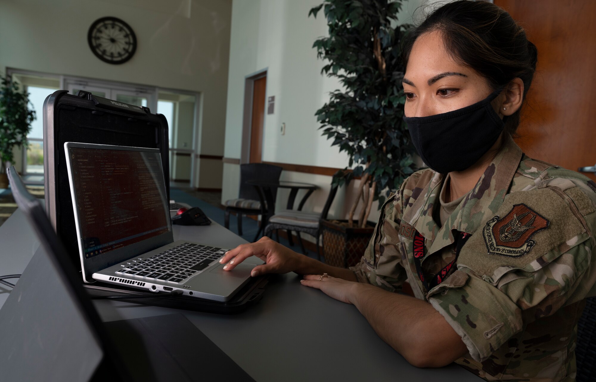 A woman wearing a facemask at a desk looks at a laptop.