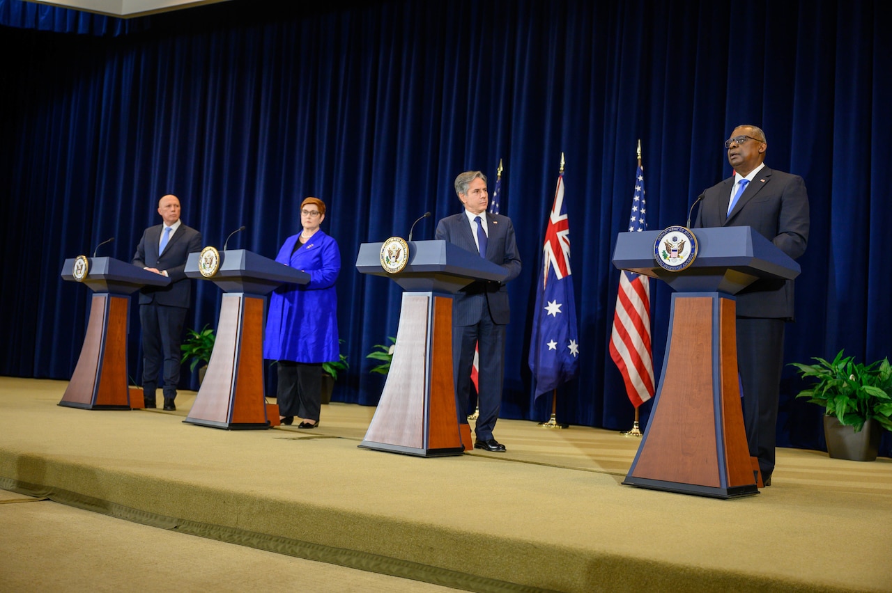 Four officials at podia with U.S. and Australian flags behind them.