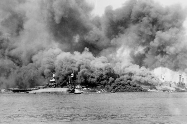 Thick, black smoke fills the sky above several burning ships; one ship capsizes.