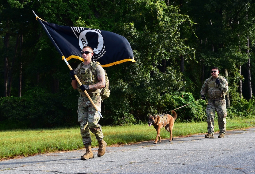 An airman carries the pow/mia flag along a rural road; a dog and another airman follow.