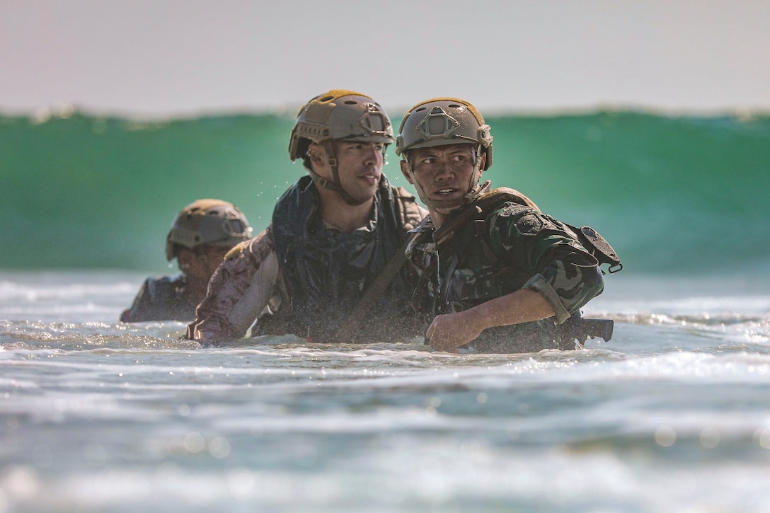 Marines stand in the beach as a wave emerges.