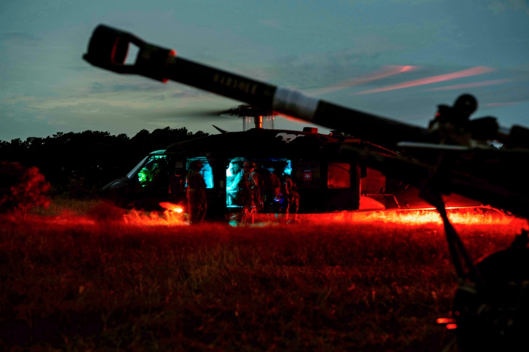 Soldiers sit in a helicopter parked in a field illuminated by colorful lights.