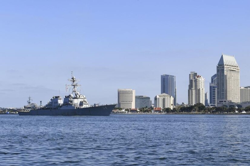A military ship steams on the water with the buildings of a city behind it.