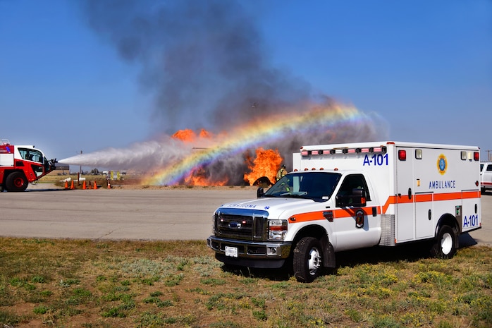 An ambulance is parked while firefighters spray water at an aircraft fire training simulator ablaze in the background.