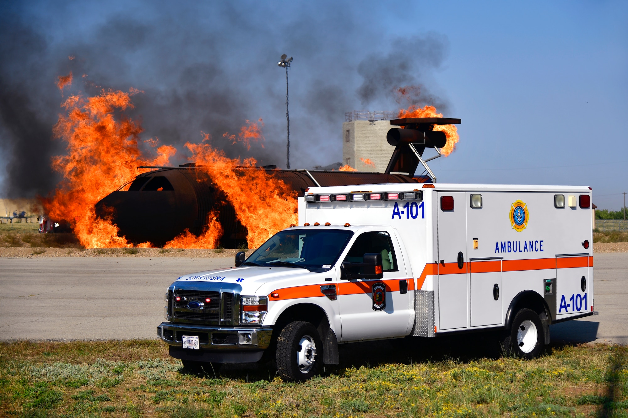 An ambulance parked while an aircraft fire training simulator is ablaze in the background.