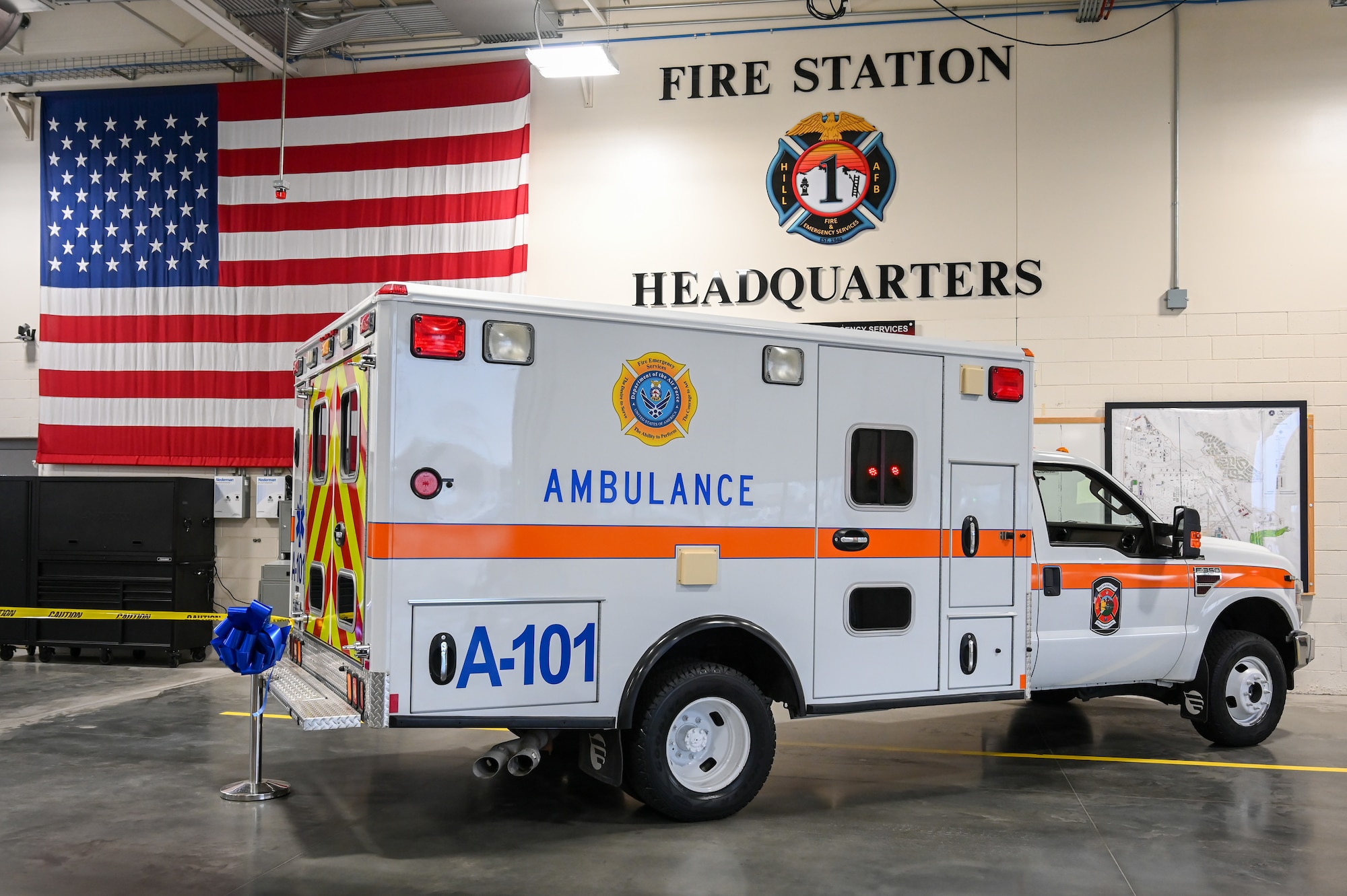 An ambulance inside the bay of the fire station with the American flag and fire station headquarters emblem on the wall behind it.