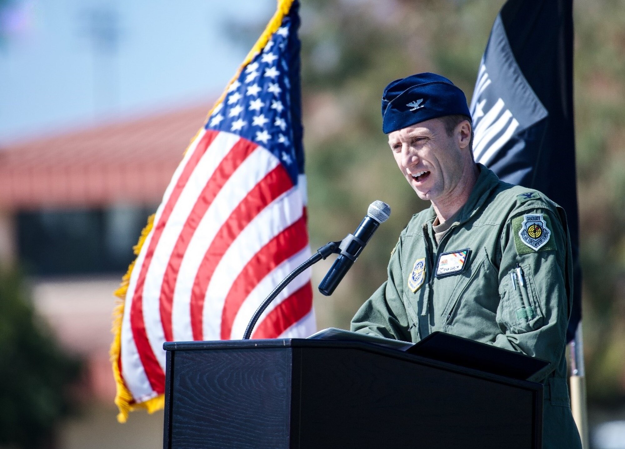 A uniformed Airman speaks into a microphone in front of the U.S. and POW/MIA flags