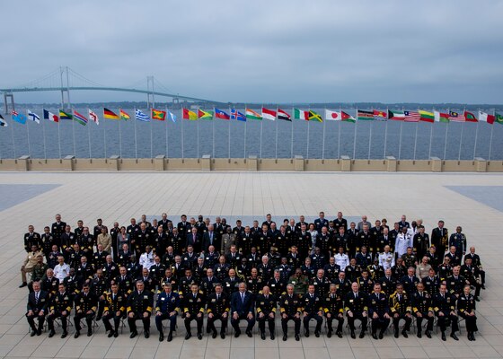 A group of people sit posed for a photo on a deck with flags streaming from the banister and the Pell Bridge in the background.