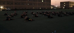 36 people lay on the ground with bent legs on a green turf field in the early morning.