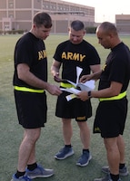 three men stand next to each other in black shorts and black shirts with yellow words and yellow reflective belts looking at groups of paper