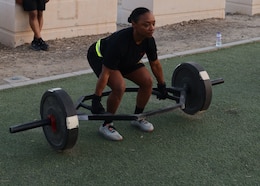 female in black shorts and shirt with yellow writing and a yellow reflective belt picks up a hex bar with weights.