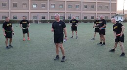 a group of men in black shorts and shirt with yellow writing stand in a circle while a man in black shorts and a blue t-shirt stands in the middle of the circle talking to them.