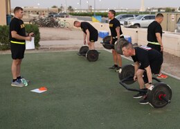 man in black shorts with a black t-shirt with yellow writing and a yellow reflective belt stands in front of three men picking up a hex bar with weights