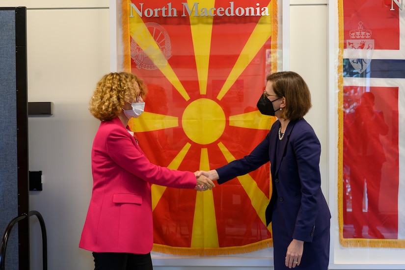 Two women shake hands in front of a yellow and red flag.