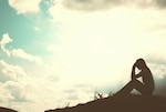 Silhouette of a person sitting on the group holding their head in their hands. Backdrop is of the blue sky.