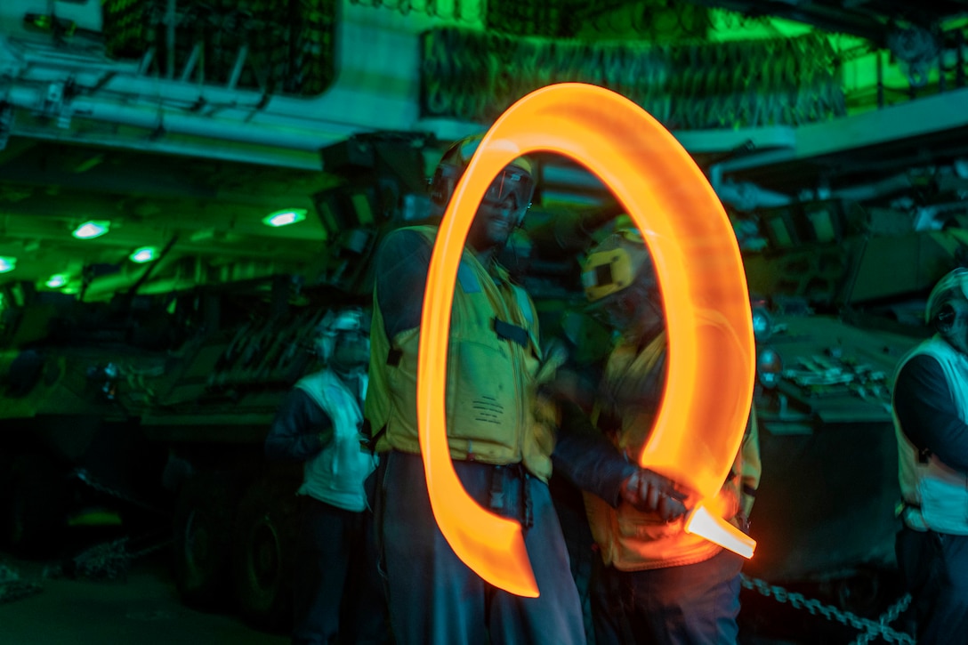 A sailor signals while standing on a ship with military vehicles parked in the background illuminated by an orange light.