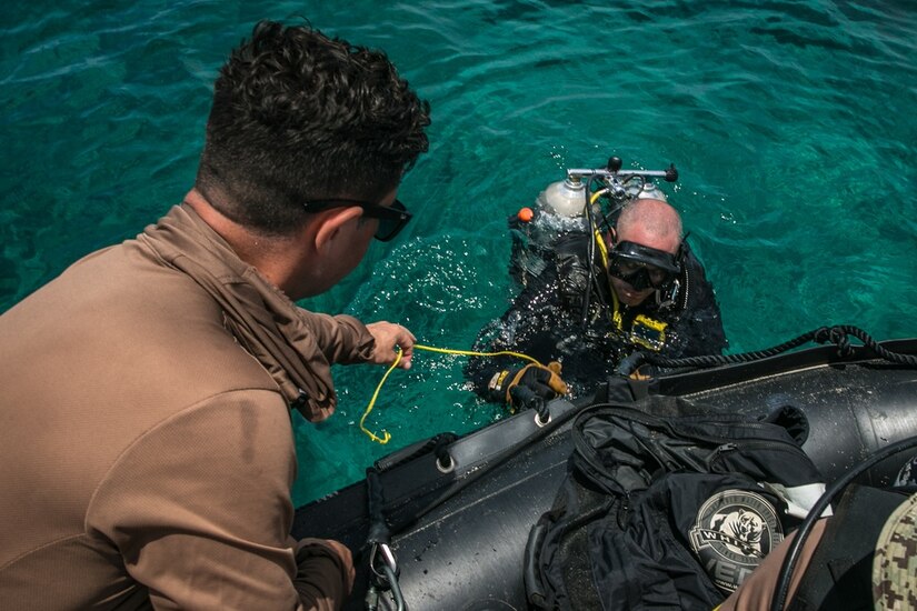 A sailor in dive gear holds onto a rubber boat as another person helps pull him in.