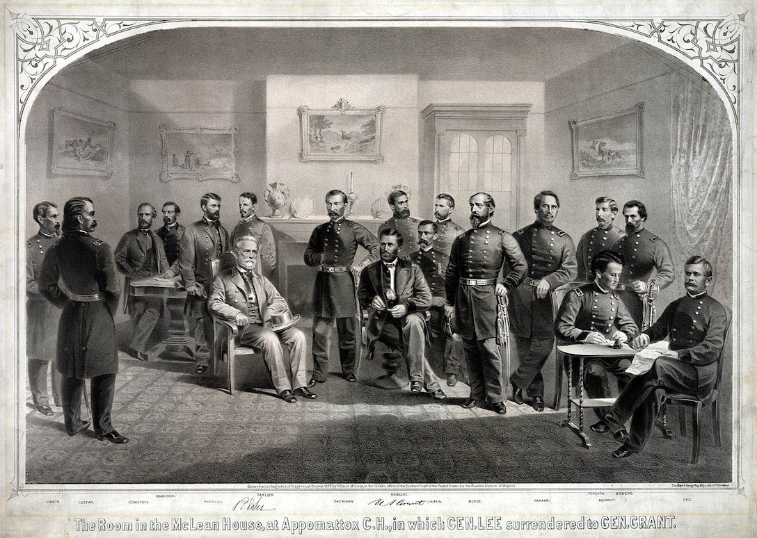 Fifteen men in military uniforms stand in a small room; two men sit in the center of the room.