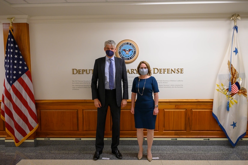 A man and a woman stand in a lobby. The signage on the wall behind them indicates that they are at the Department of Defense.