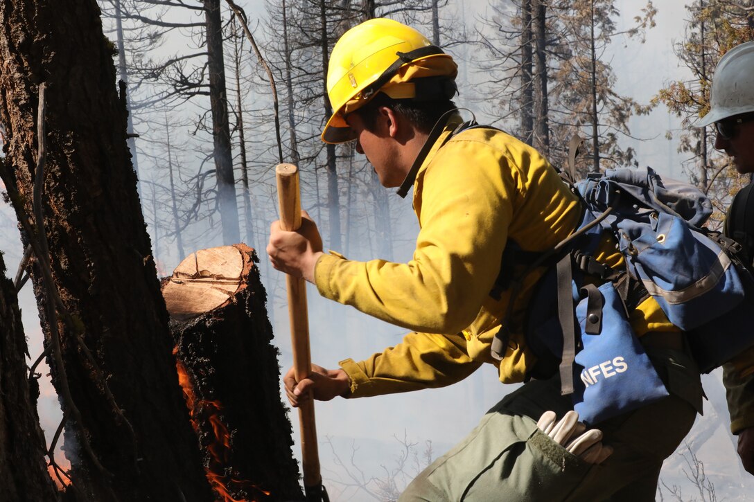 A soldier dressed in firefighting gear uses a tool to help stop the spread of a fire.