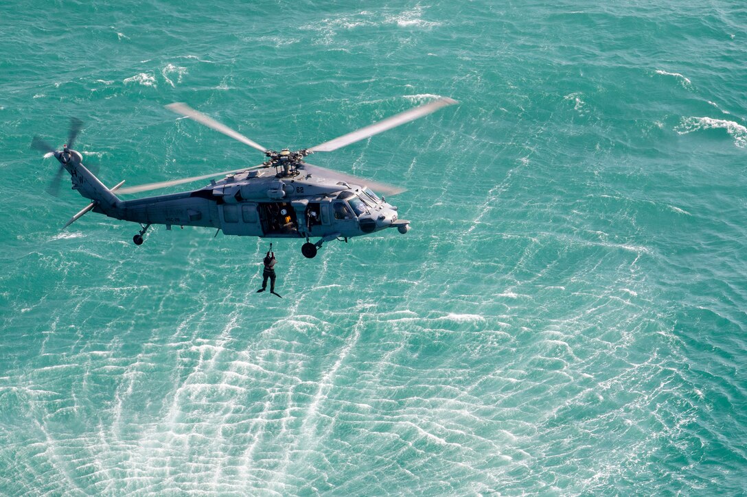 A sailor holds onto a rope attached to an airborne helicopter flying over a body of water.