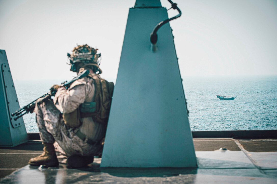 A Marine kneels on the deck of a ship as another vessel sails in the distance.