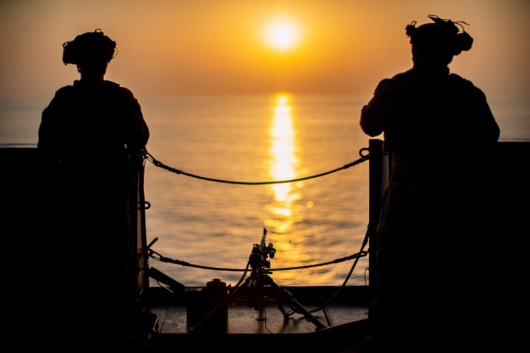 Marines looking at the sun stand on a ship transiting a body of water as shown in silhouette.