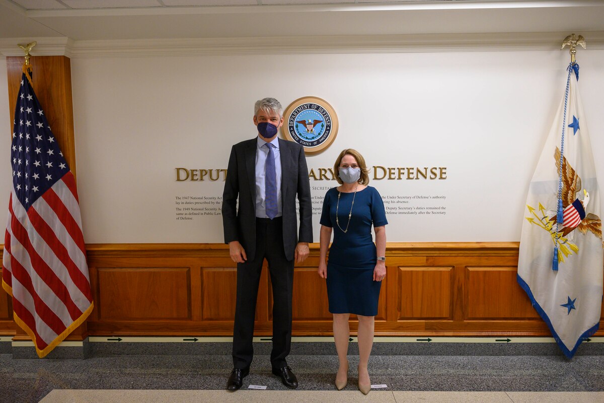 A man and a woman stand in a lobby. The signage on the wall behind them indicates that they are at the Department of Defense.