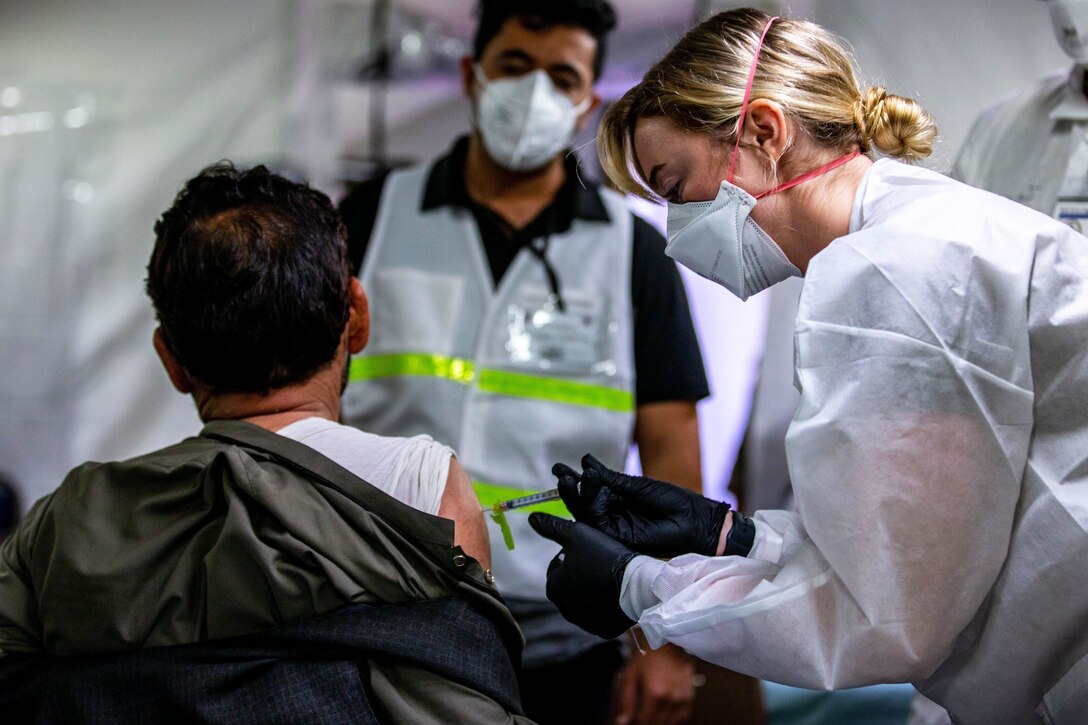 A woman wearing a face mask, gloves and medical gown bends down while holding a syringe to give a vaccine to a man.