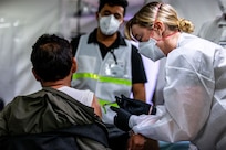 A woman wearing a face mask, gloves and medical gown bends down while holding a syringe to give a vaccine to a man.