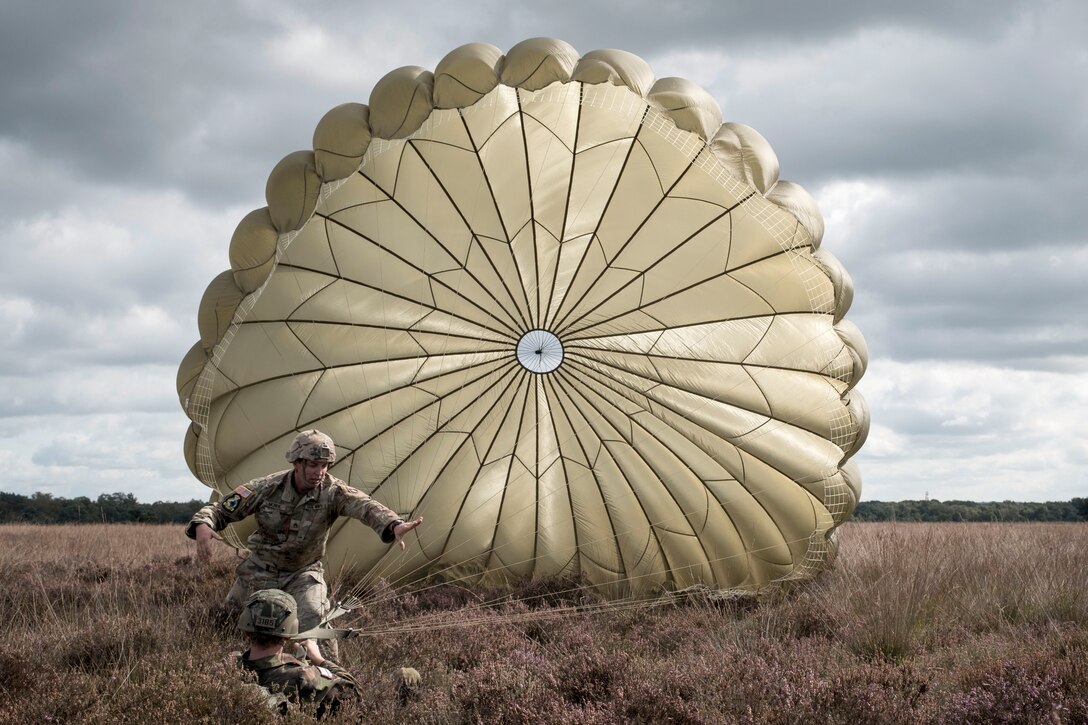 A soldier standing in a field works with another on the ground attached to an extended parachute canopy behind them