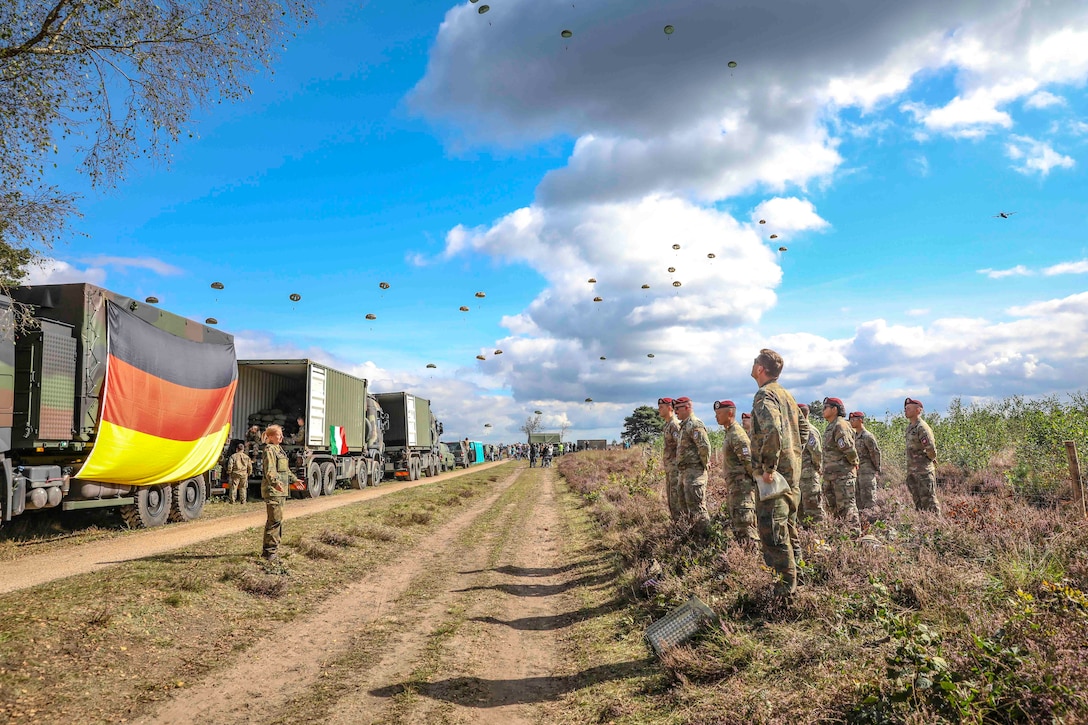 Soldiers stand alongside a dirt road as others descend in the sky wearing parachutes.