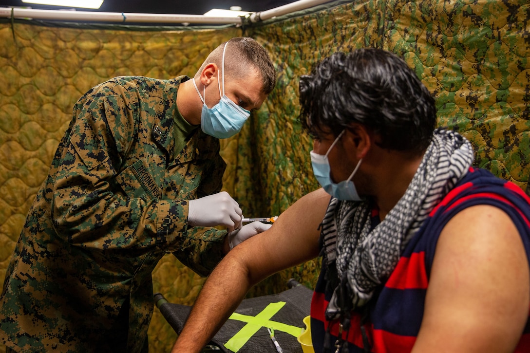 A sailor wearing a face mask and glove bends over while holding a syringe to give a  vaccine to a man wearing a face mask sitting down.