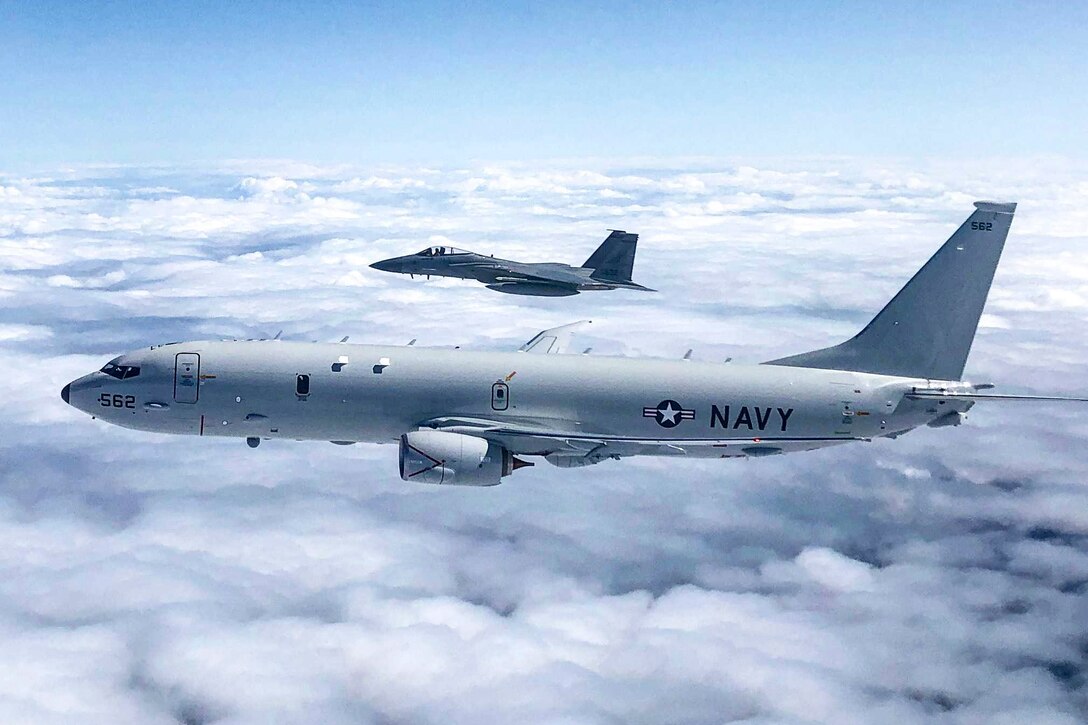 A Navy aircraft flies next to an Air Force jet above clouds.