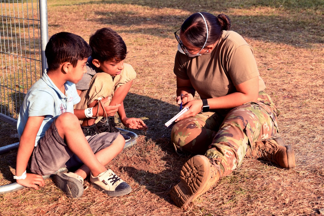 A service member sits on the ground with two boys and points at a paper.