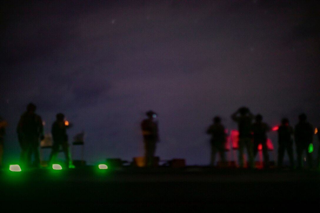 Marines fire weapons on a ship at night illuminated by red and green lights.
