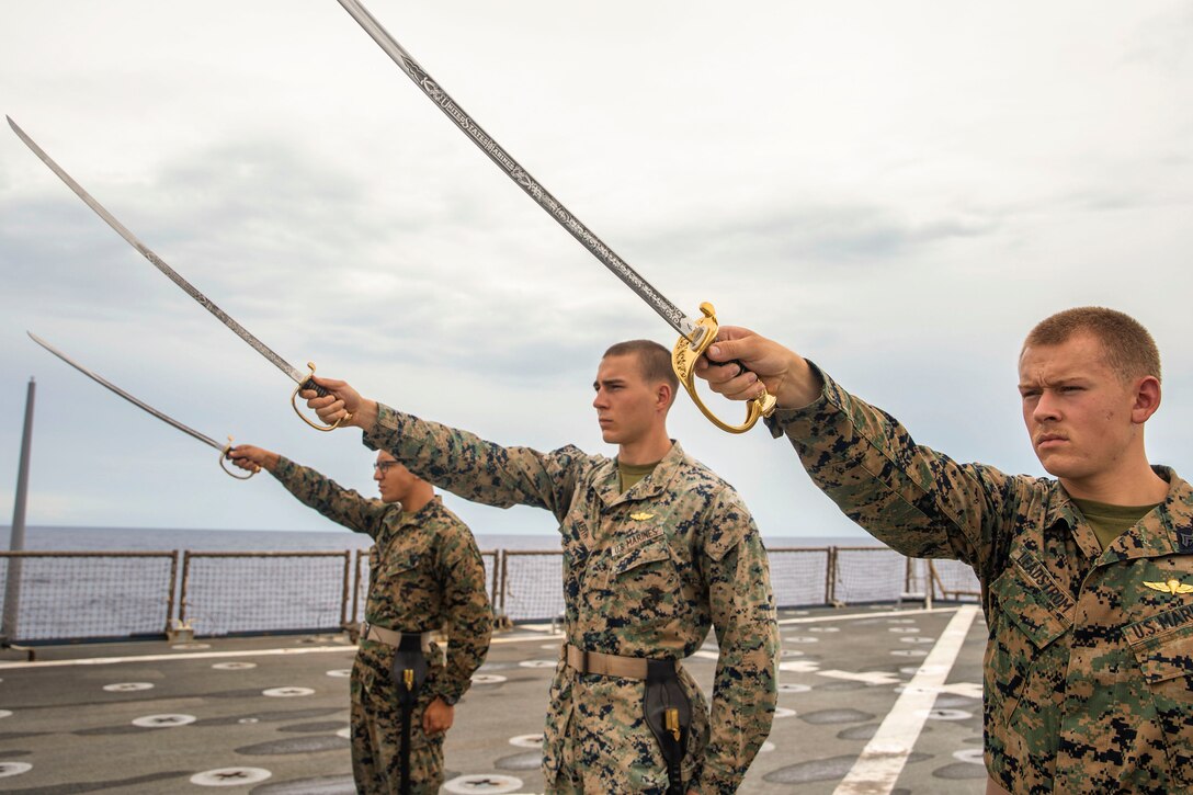 Marines draw swords while standing on a ship at sea.