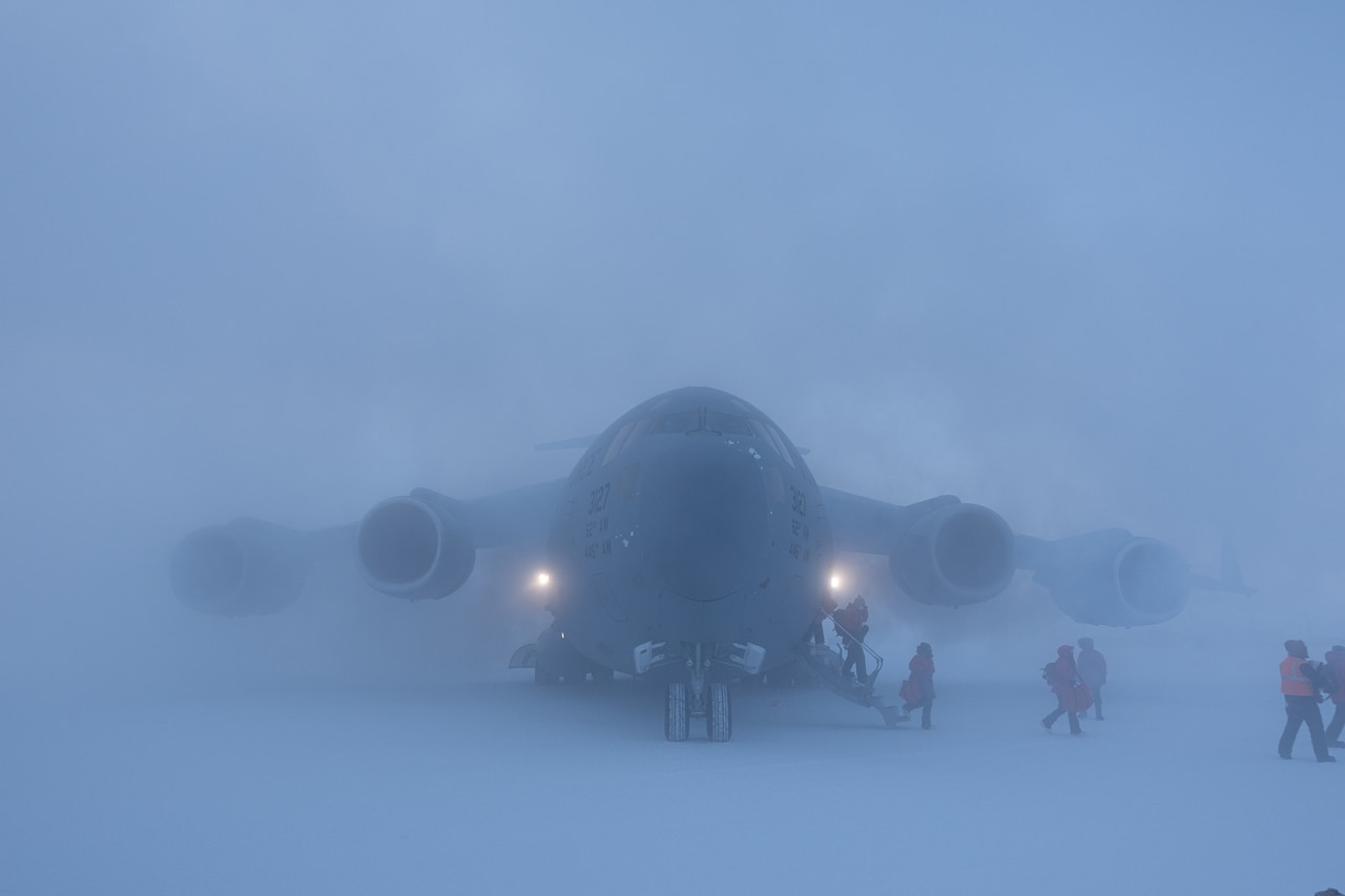 A large aircraft is barely seen through snow mist as people walk to the aircraft.