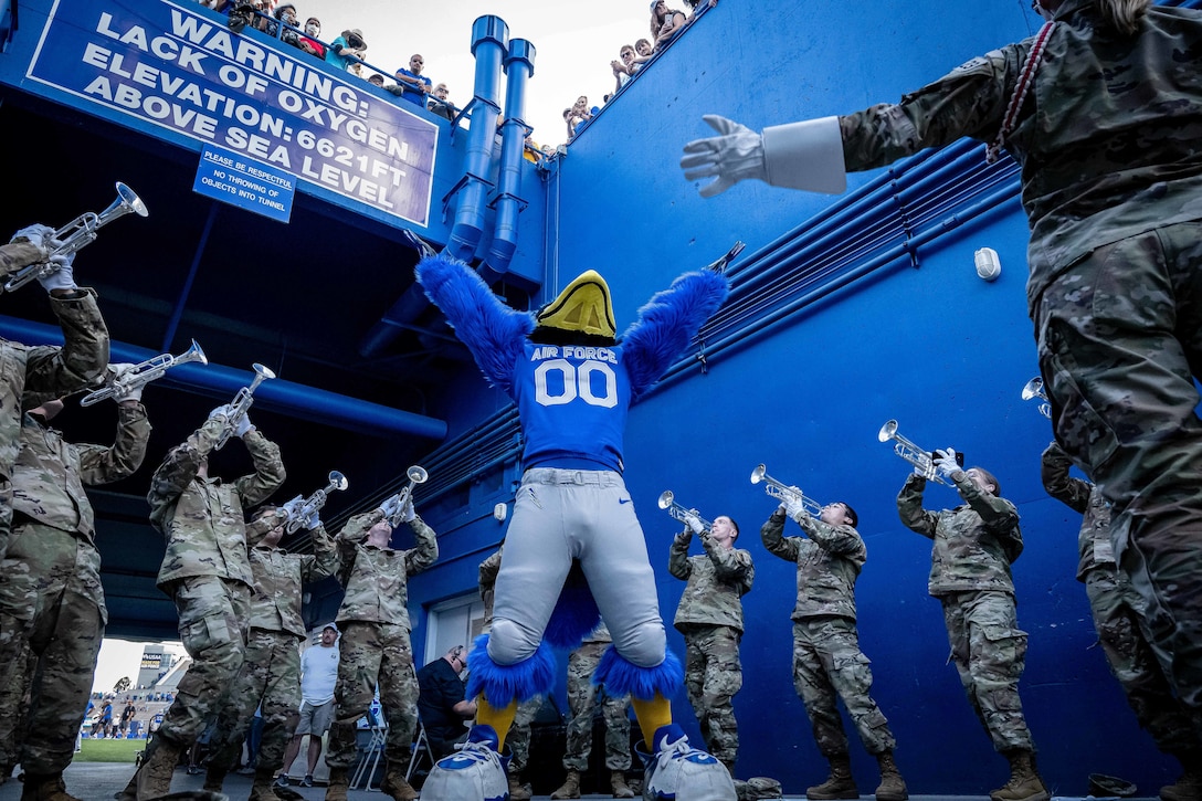 A person in a bird costume raises his hands in the air as a group of airmen play instruments in a stadium.