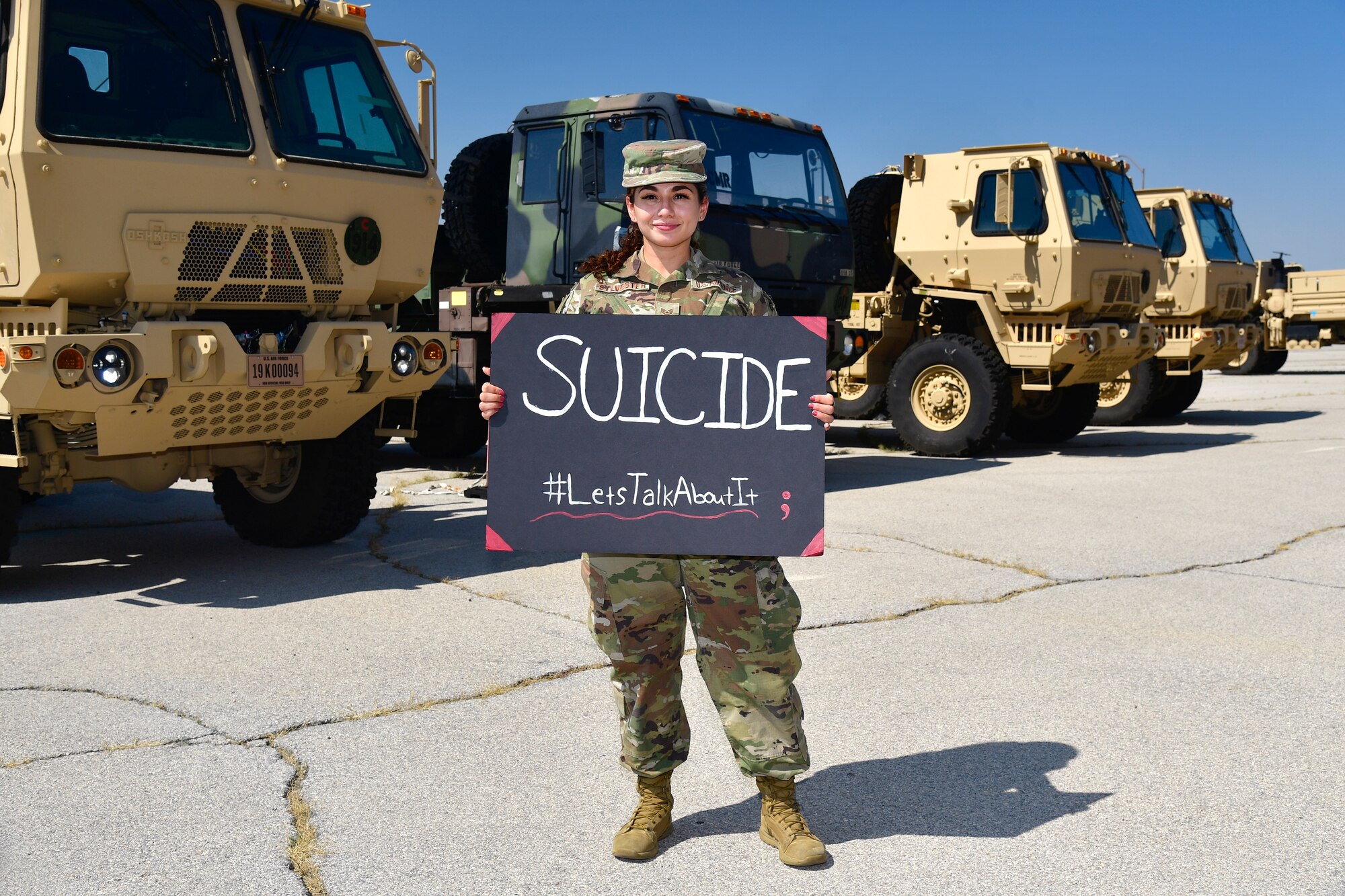 Staff Sgt. Gabriela Sylvester holding a sign that reads “Suicide #LetsTalkAboutIt” with military trucks in the background.