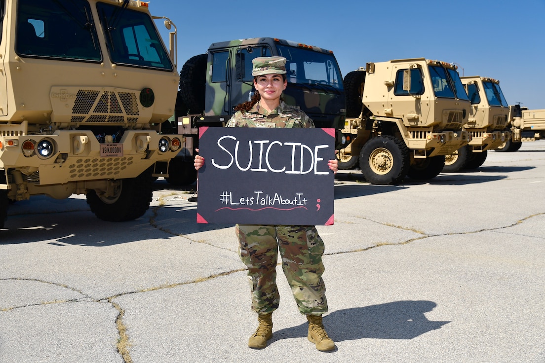 Staff Sgt. Gabriela Sylvester holding a sign that reads “Suicide #LetsTalkAboutIt” with military trucks in the background.